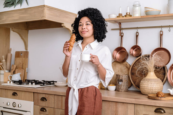 africká americká žena stojící v blízkosti dřevěné skříně s pultem držící šálek horkého bylinného čaje a jíst čerstvý francouzský croissant k snídani doma kuchyně s útulným interiérem.. - Fotografie, Obrázek