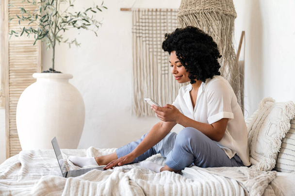 Iloinen afro amerikkalainen nainen työskentelee kannettavan ja tarkistaa online app hänen moderni älypuhelin. Freelance nainen istuu sängyllä viihtyisässä makuuhuoneessa, kirjallisesti postitse blogiin. Etätyö kodin käsitteistä - Valokuva, kuva