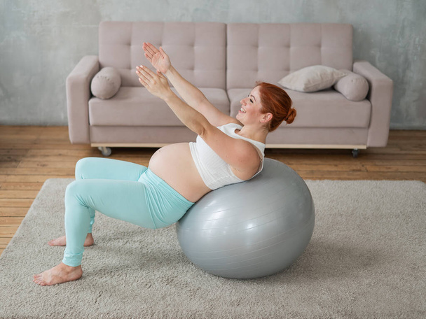 Pregnant red-haired woman doing exercises on fitness ball at home - Φωτογραφία, εικόνα