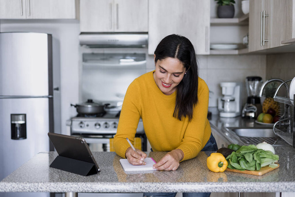 Latynoska dorosła kobieta gotowanie i pisanie receptury w notatniku w kuchni w domu w Meksyku, Latynoska kobieta przygotowuje meksykańskie jedzenie w Ameryce Łacińskiej - Zdjęcie, obraz