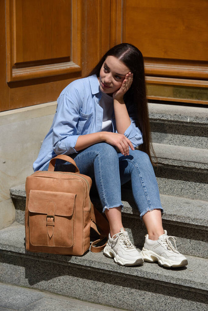 Красивая молодая девушка в синих джинсах, рубашке и белой футболке позирует с коричневым рюкзаком из кожи. Фото с улицы - Фото, изображение