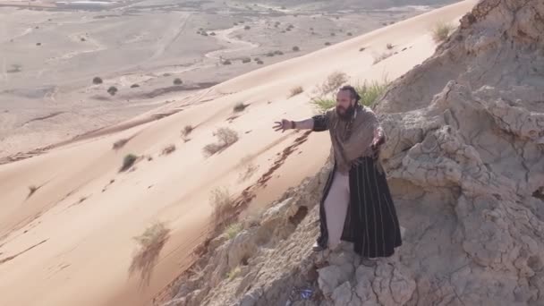 Um homem com um cajado na mão e as roupas de um xamã está em uma rocha entre as areias do deserto e danças. Movimento lento, drone aéreo - Filmagem, Vídeo