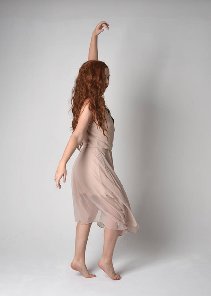 Full length πορτρέτο του όμορφου γυναικείου μοντέλου με μακριά καστανά μαλλιά φορώντας ένα κρεμώδες ροζ φόρεμα. χαριτωμένη χορευτική στάση, με χειρονομιακά χέρια απομονώνονται σε λευκό φόντο στούντιο. - Φωτογραφία, εικόνα