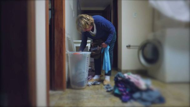 Uma mulher idosa sincera a fazer tarefas de lavandaria em casa. Cena doméstica de uma senhora mais velha removendo roupas da cesta - Foto, Imagem