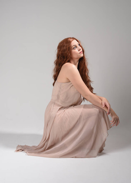 Portrait pleine longueur d'une belle mannequin brune portant une robe rose. pose assise gracieuse, agenouillée sur le sol mains gestuelles. prise de vue en angle bas, isolée sur fond de studio blanc. - Photo, image