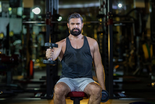 Kaukázusi szakállas izmos sport férfi gyakorolja súlyzós edzés súlyzót bicepsz és tricepsz izom belsejében tornaterem sötét háttér és világos füst világít gyakorlása és edzés használata - Fotó, kép