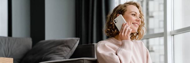 Белая женщина улыбается и разговаривает по мобильному телефону, сидя дома на полу - Фото, изображение