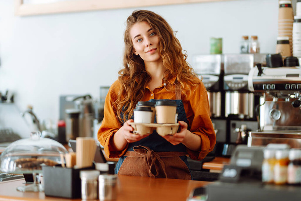Lachende vrouwelijke barista die koffie geeft om naar klanten te gaan. Een krullende koffieshop eigenaar werkt achter de toonbank. Bedrijfsconcept. meeneemvoedsel - Foto, afbeelding