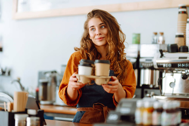 Smiling female barista giving coffee to go to customers. A curly-haired coffee shop owner works behind the counter. Business concept. takeaway food - Фото, изображение