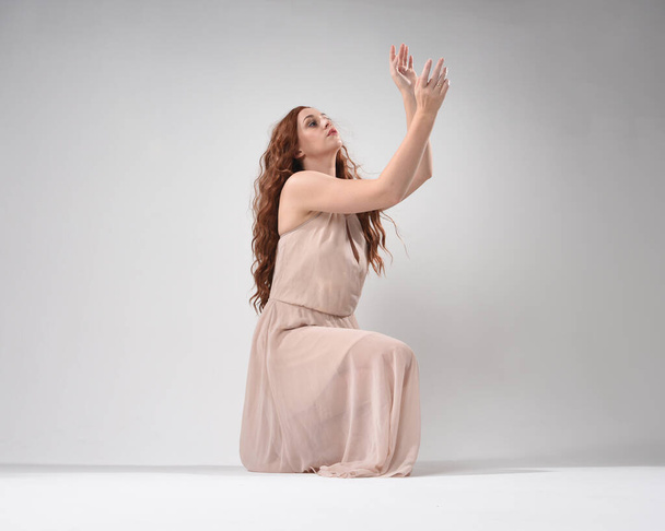 Volledig portret van een mooi brunette model in een roze jurk. sierlijke zittende houding, knielend op de vloer gebaren handen. opgenomen vanuit lage hoek perspectief, geïsoleerd op witte studio achtergrond. - Foto, afbeelding
