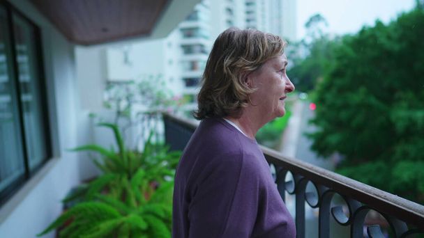 Eine nachdenkliche Seniorin blickt an einem regnerischen Nachmittag vom Balkon ihrer Wohnung. Eine nachdenkliche ältere Dame in den Siebzigern betrachtet das Leben und starrt auf den fallenden Regen - Foto, Bild