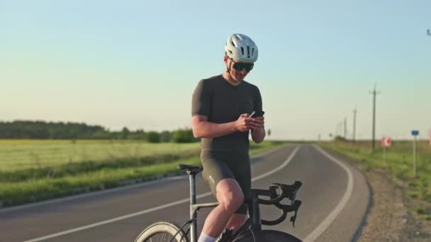 Добре побудований здоровий чоловік у спортивному одязі, використовуючи розумний пристрій, тримаючи чорний велосипед під час короткої перерви на дорозі. Задоволений гонщик перевіряє дані продуктивності через додаток велосипедного комп'ютера по телефону. - Кадри, відео