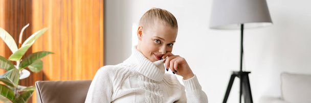 Smiling young white woman with blonde hair wearing sweater sitting on a chair with laptop computer at home - Φωτογραφία, εικόνα