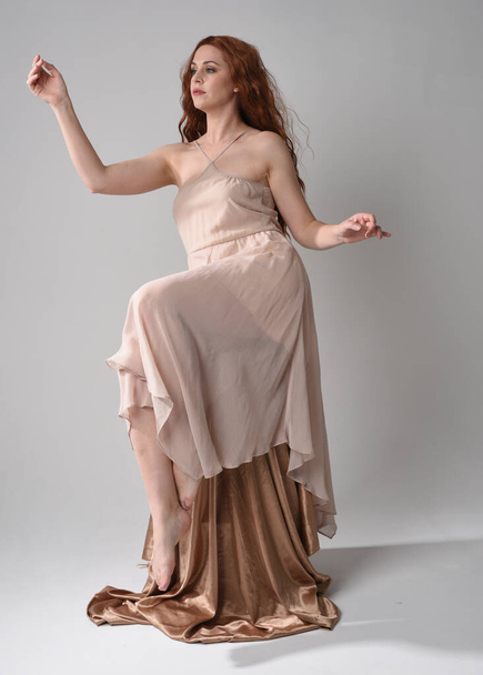 Portrait pleine longueur d'un beau modèle féminin avec de longs cheveux bruns portant une robe rose crème. gracieuse pose assise isolée sur fond de studio blanc. - Photo, image
