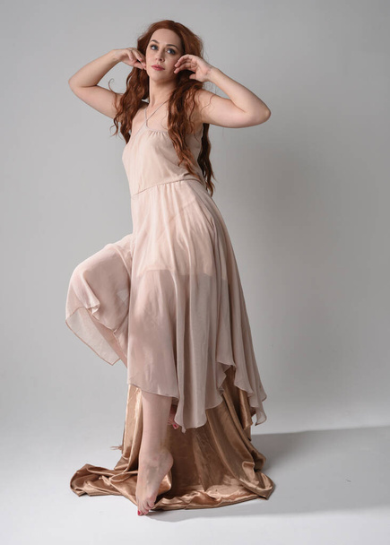 Full length portrait of beautiful  female model with long  brunette hair wearing a creamy pink gown dress. graceful sitting pose isolated on white studio background. - Φωτογραφία, εικόνα