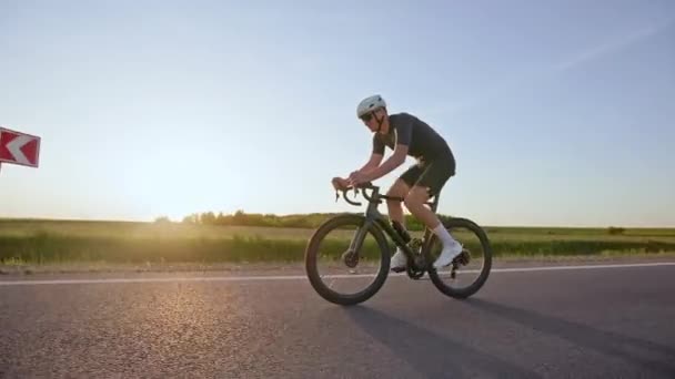Volledig uitgeruste racer op racefiets uitvoeren van oefening rit met hoge snelheden langs de weg op veld achtergrond. Sportieve mannelijke fietser in strakke kleding die zich aan het drukke trainingsschema houdt bij zonsondergang. - Video