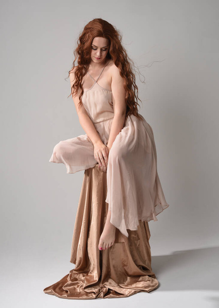 Portrait pleine longueur d'un beau modèle féminin avec de longs cheveux bruns portant une robe rose crème. gracieuse pose assise isolée sur fond de studio blanc. - Photo, image