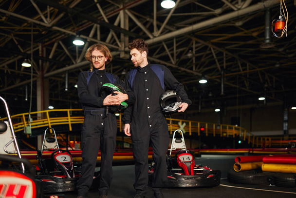 two kart racers standing near racing cars and holding helmets, male drivers in kart circuit - Fotó, kép
