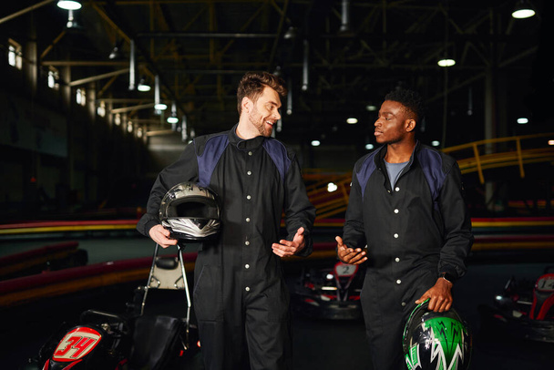 happy multicultural men chatting inside of kart circuit, kart racers walking and holding helmets - Fotografie, Obrázek