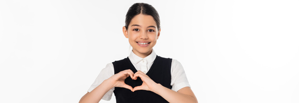fröhliches Schulmädchen zeigt Herzzeichen mit Händen und blickt vereinzelt in die Kamera auf weißem Banner - Foto, Bild