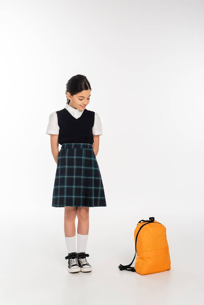 χαρούμενο κορίτσι με σχολική στολή στέκεται και κοιτάζει το σακίδιο στο λευκό φόντο, πίσω στο σχολείο - Φωτογραφία, εικόνα
