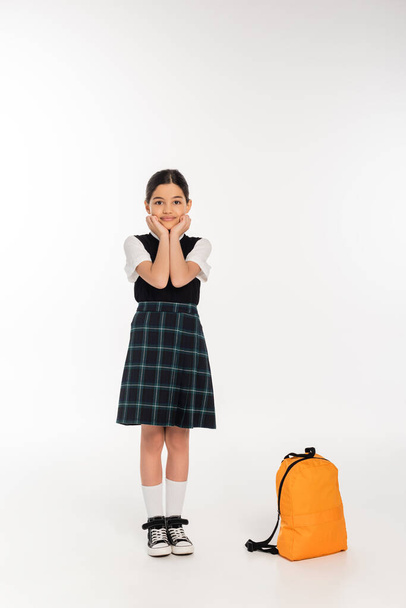 happy girl in school uniform standing near backpack on white background, back to school, full length - Photo, Image