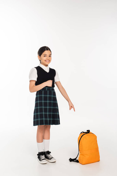 alegre chica en uniforme de la escuela de pie y apuntando a la mochila sobre fondo blanco, longitud completa - Foto, imagen