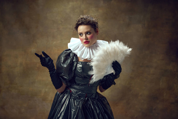 Φωτογράφηση της γυναίκας σε φόρεμα μπάλα από σακούλες σκουπιδιών και ανεμιστήρα στο χέρι πάνω από vintage φόντο. Πορτραίτο των αριστοκρατικών γυναικών. Έννοια περιβαλλοντικών θεμάτων, ρετρό, ρετρό, στυλ, μόδα. - Φωτογραφία, εικόνα