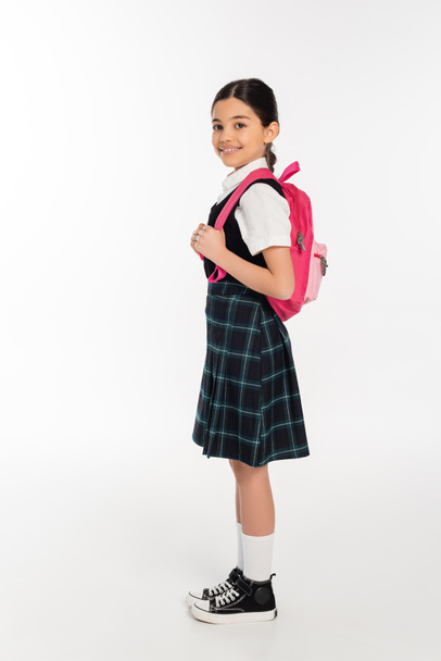 plná délka, šťastná školačka stojící v uniformě s batohem na bílém, připravená na nový školní rok - Fotografie, Obrázek