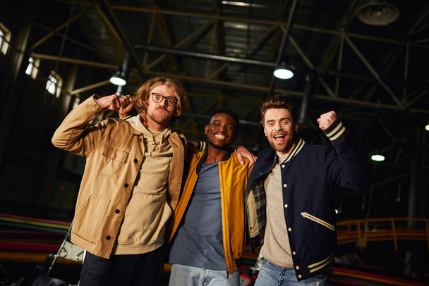 three interracial male friends smiling and standing near racing cars inside of indoor karting track - Photo, Image