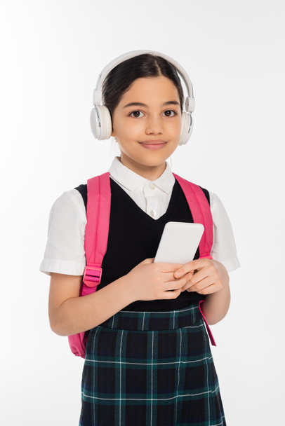 цифровий вік, радісна школярка в бездротових навушниках тримає смартфон ізольовано на білому, студент - Фото, зображення