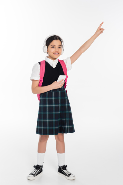 digital age, cheerful schoolgirl in wireless headphones holding smartphone on white, pointing away - Foto, afbeelding