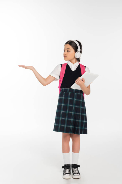 školačka v bezdrátových sluchátkách držící digitální tablet na bílém, digitálním věku, ukazující rukou - Fotografie, Obrázek