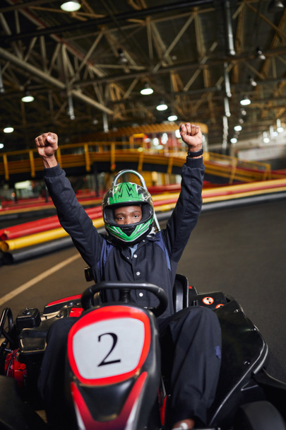go kart speed drive, thrilled african american driver in helmet raising hands and winning kart race - Foto, afbeelding