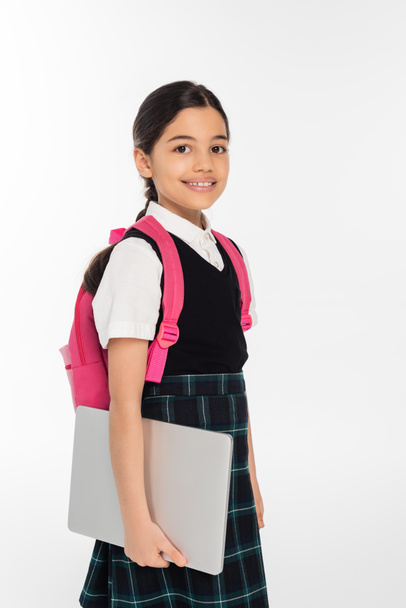 pozytywna uczennica trzymająca laptopa i patrząca w kamerę, dziewczyna w szkolnym mundurze, odizolowana na białym - Zdjęcie, obraz