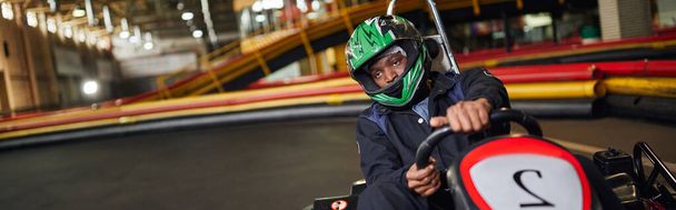サーキット上のヘルメットのアフリカ系アメリカ人ドライバー, モータースポーツのコンセプトをカート, 水平バナー - 写真・画像