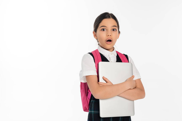 σοκαρισμένη μαθήτρια κρατώντας φορητό υπολογιστή και κοιτάζοντας κάμερα, κορίτσι με σχολική στολή, απομονωμένη σε λευκό - Φωτογραφία, εικόνα