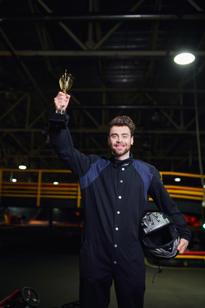veselý řidič ve sportovním oblečení drží zlatý pohár a stojí jako vítěz motokáry závod, trofej - Fotografie, Obrázek