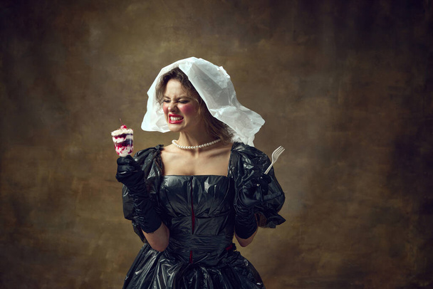 Księżniczko. Portret kobiety ubranej w polietylenową białą czapkę i czarną sukienkę z worków na śmieci zgrzytających zębami i trzymającej pyszny deser. Pojęcie kwestii środowiskowych, recyklingu, stylu, mody. - Zdjęcie, obraz
