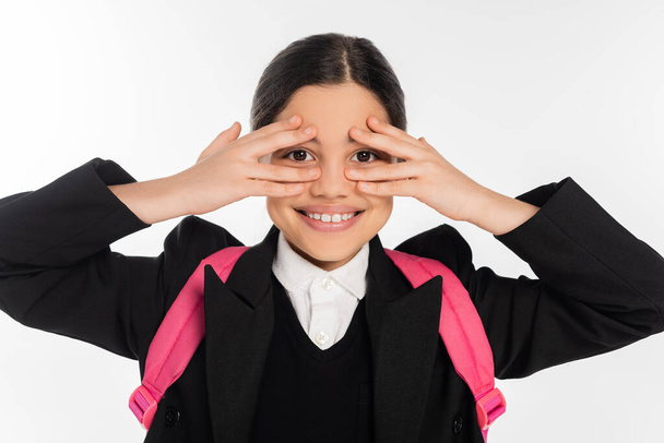 happy schoolgirl in uniform covering eyes isolated on white, student life, looking at camera - Photo, Image