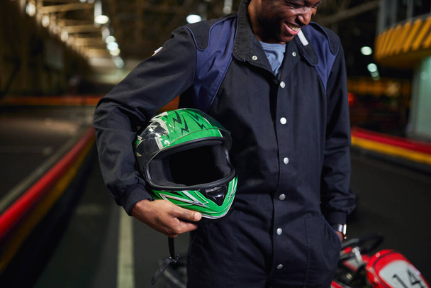 ヘルメットを保持し,カートのトラックサーキットに立っているスポーツウェアの陽気なアフリカ系アメリカ人男性 - 写真・画像