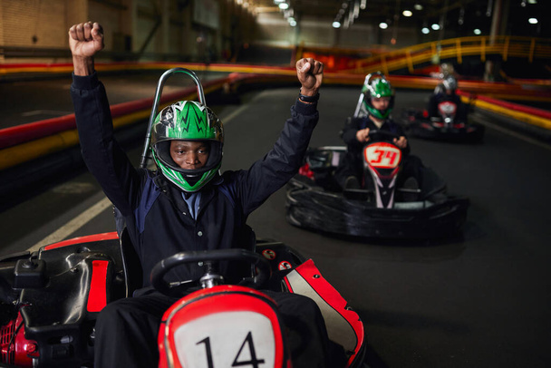aufgeregter afrikanisch-amerikanischer Go-Kart Racer im Helm hebt die Hände und gewinnt Rennen auf der Rennstrecke, Go-Kart - Foto, Bild
