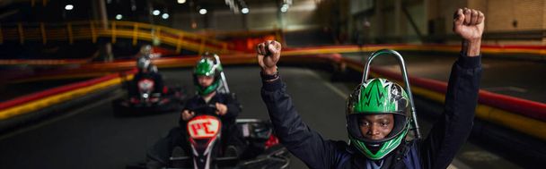 vzrušený africký americký go vozík řidič v helmě zvedání rukou a vítězné závod, go-kart prapor - Fotografie, Obrázek