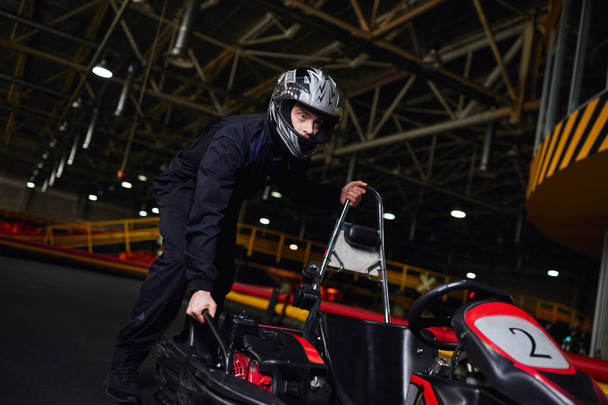 automovilismo y velocidad, conductor de kart enfocado en casco y ropa deportiva empujando karts en circuito - Foto, Imagen