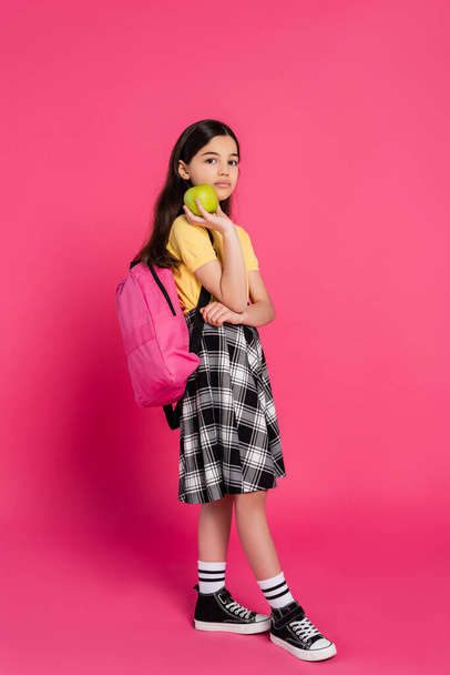 full length, brunette schoolgirl standing with backpack and holding green apple, pink background - Photo, Image