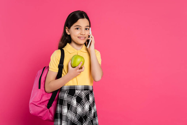 gelukkig schoolmeisje staan met rugzak, met appel en praten op smartphone, telefoongesprek - Foto, afbeelding
