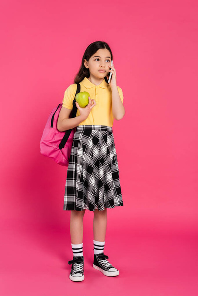 brunette schoolgirl talking on smartphone on pink background, phone call, apple and backpack - Photo, Image