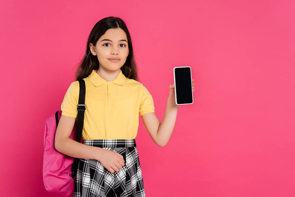 brunette écolière tenant smartphone avec écran vide et regardant la caméra sur fond rose - Photo, image
