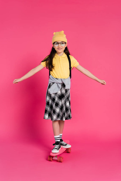 happy schoolgirl in beanie hat and glasses riding penny board on pink background, stylish look - Photo, Image