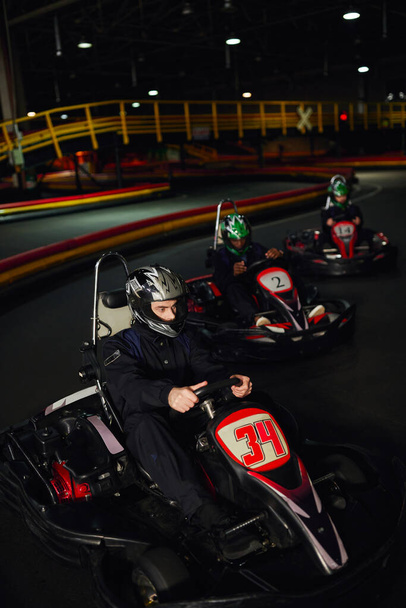focused man driving go kart near diverse drivers in helmets on indoor circuit, adrenaline and sport - Photo, Image